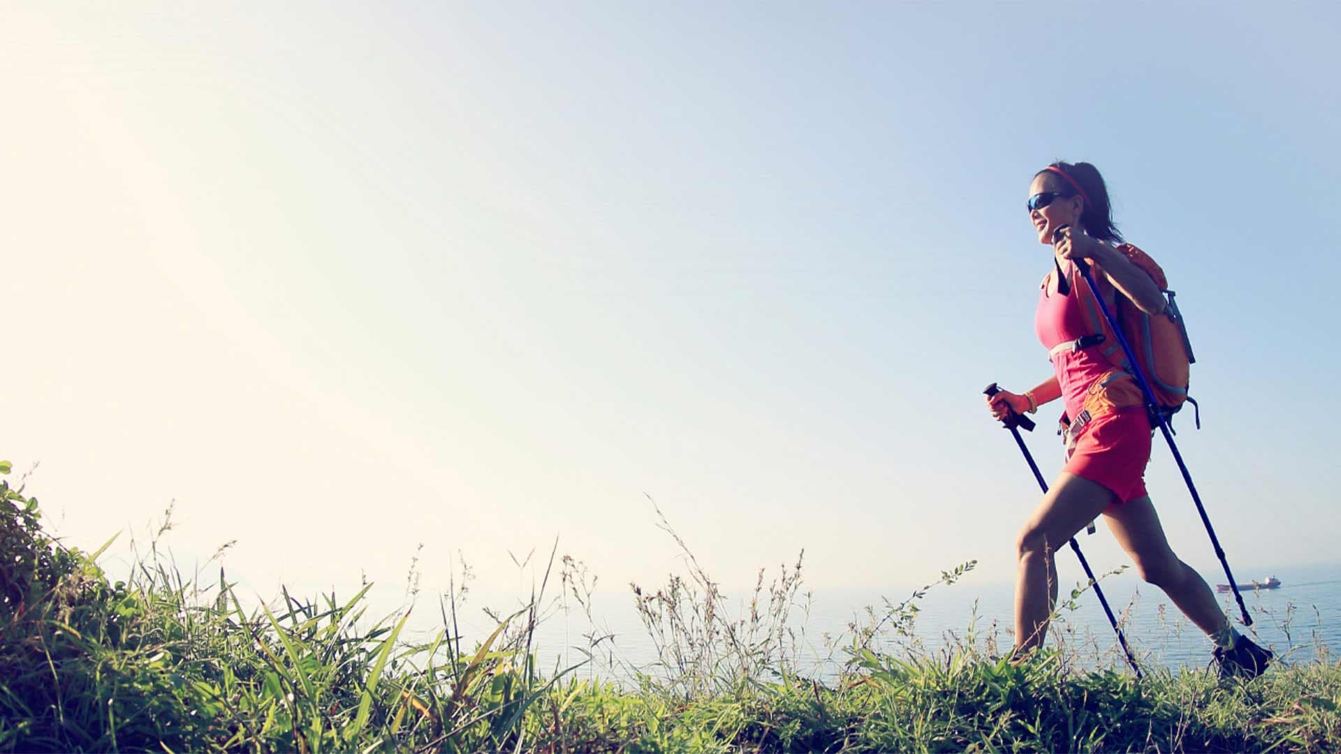 A woman walking through the grass with hiking poles, with a body of water behind her.