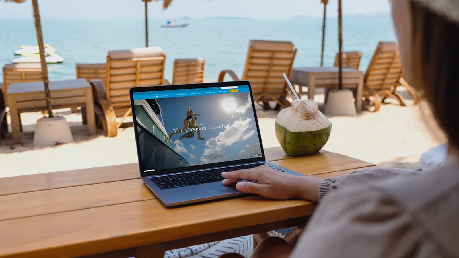 A person sits at a table on the beach looking at a Cayman Islands website on a laptop