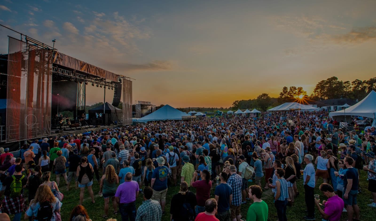 Music fans at an outdoor concert at sunset with a stage in the background