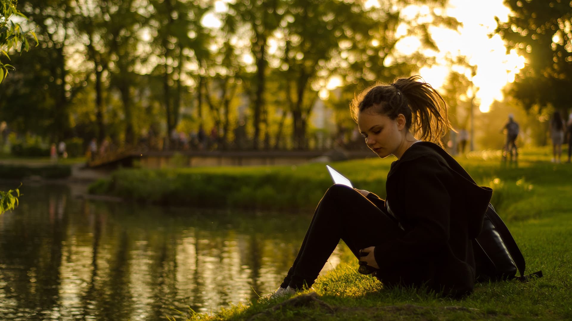 A student sits on the edge of a pond doing school work