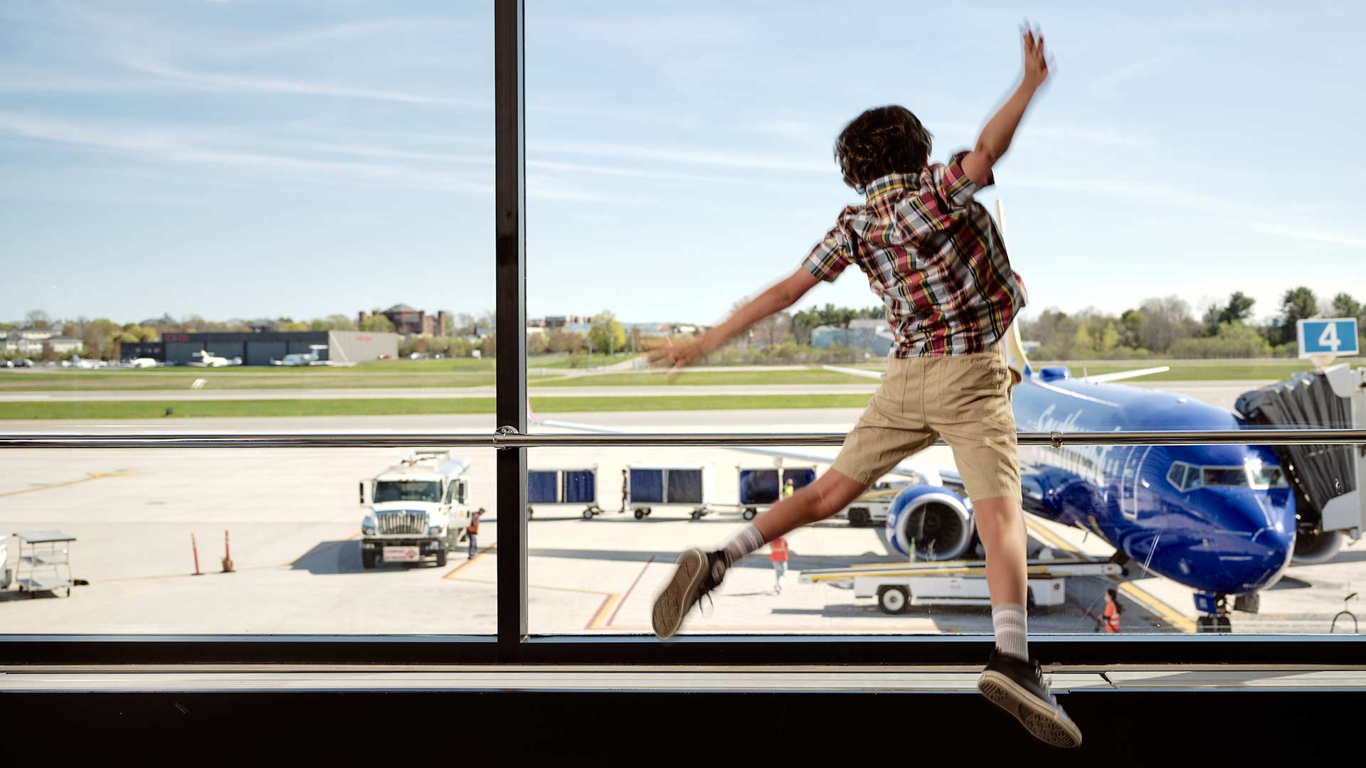 A boy leaps in front of a window watching an airplane on the tarmac.