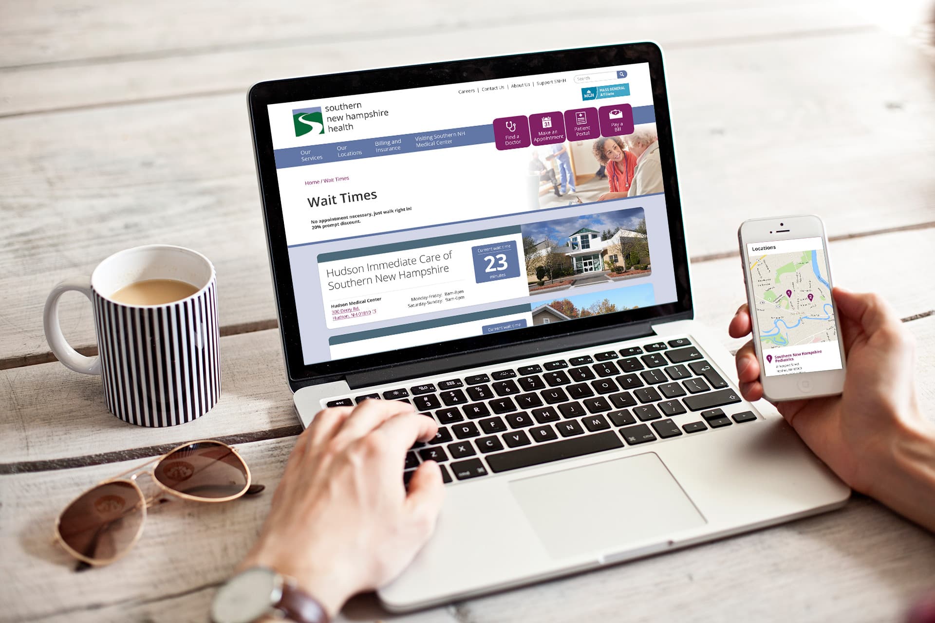 A laptop displaying the Southern New Hampshire Healthcare website sits on a table. Next to it, an iPhone with a map, sunglasses, and a mug are placed on the surface.