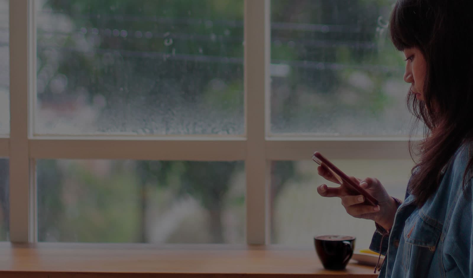 A girl using her phone while standing by a window on a rainy day
